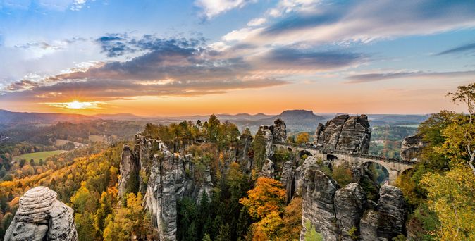 Eine spektakuläre Felsformation und die Basteibrücke in der Sächsischen Schweiz, umgeben von einem dichten Wald, unter einem bewölkten Himmel.