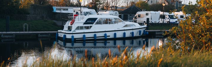 Ein kleines, weißes Motorboot mit dem Schriftzug "CARRICKCRAFT" und blauen Akzenten, das auf einem ruhigen Fluss oder See fährt. Im Hintergrund ist eine bewaldete Uferlandschaft mit weiteren Booten zu sehen.