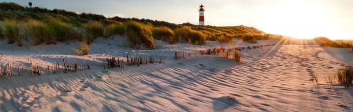 Leuchtturm auf einer Düne im Sonnenuntergang mit langen Schatten auf dem fein strukturierten Sand.