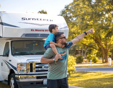 Ein Mann mit Sonnenbrille hat ein Kind auf seinen Schultern, das in die Ferne zeigt, während sie vor einem weißen Sunseeker El Monte RV Wohnmobil stehen, umgeben von Bäumen.