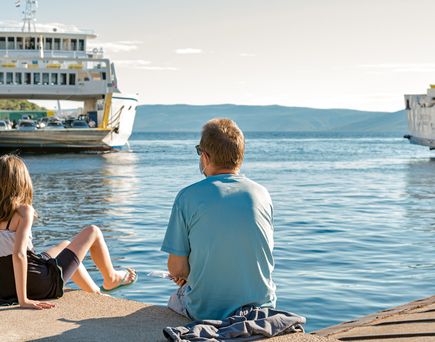 Zwei Kinder und eine erwachsene Person sitzen auf einem Bootssteg und blicken auf ein großes Schiff im Hafen. Der Himmel ist blau.