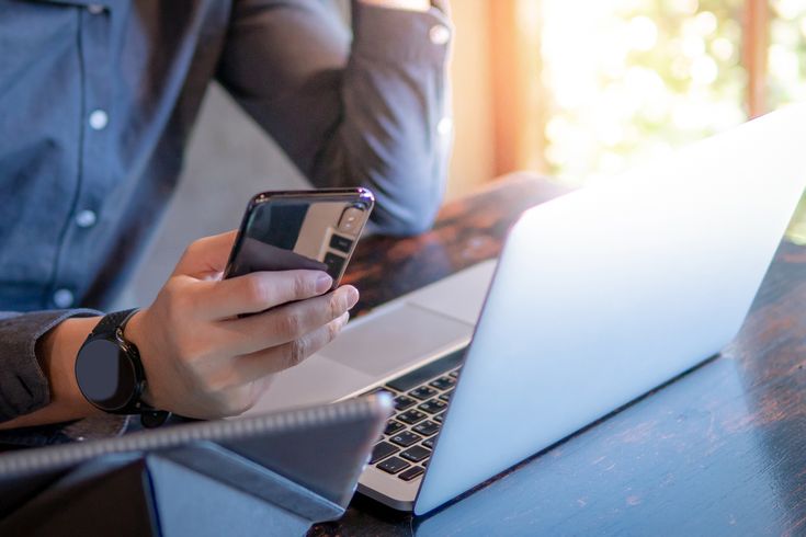 Geschäftsperson arbeitet mit Laptop und Smartphone an einem sonnendurchfluteten Tisch, moderne Arbeitsumgebung.