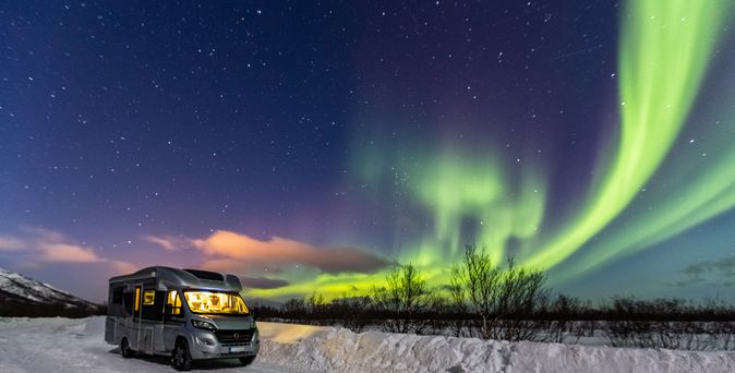 Ein Wohnmobil steht in der Nacht auf einem schneebedeckten Feld, beleuchtet von warmem Licht, das eine gemütliche Atmosphäre unter den tanzenden Nordlichtern schafft.