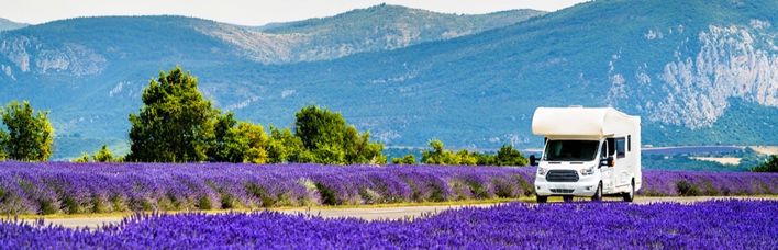 Ein weißer Campervan parkt am Rand eines blühenden Lavendelfeldes mit lila Blüten und lila Bergen im Hintergrund.