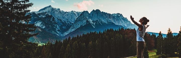 Eine Person mit ausgestreckten Armen steht auf einem Bergweg und blickt auf eine weite Gebirgslandschaft bei Garmisch-Partenkirchen, Bayern.