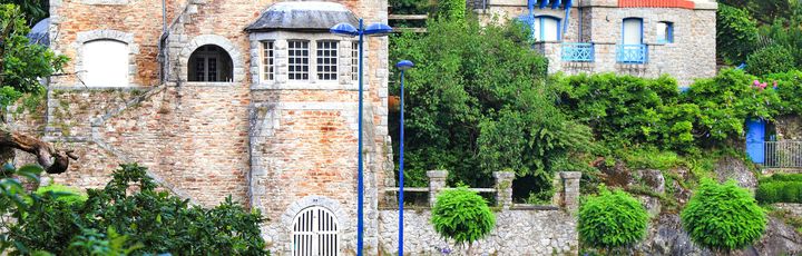 Ein charmantes, altes Steinhaus mit einer traditionellen Architektur, umgeben von grünen Pflanzen und einem blauen Himmel. Das Haus wirkt sehr malerisch.