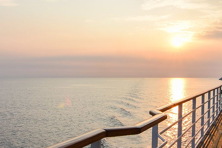 Sonnenuntergang auf dem Meer, aufgenommen vom Außendeck eines Kreuzfahrtschiffes mit hölzernem Boden und Geländer.
