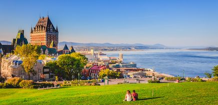 Stadtansicht der Altstadt von Quebec mit Gebäuden, grünem Gras vor dem Sankt-Lorenz-Strom und blauem Himmel an einem Sommerabend. Ein Pärchen sitz auf der grünen Wiese, mit Blick auf Gebäude und Fluss. Blauer Himmel.