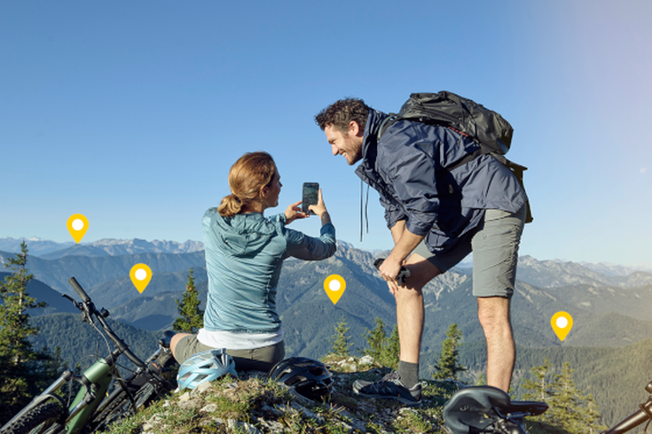 Reisepaar mit Mountainbikes genießt den Panoramablick von einem alpinen Gipfel und hält den Moment mit dem Smartphone fest.