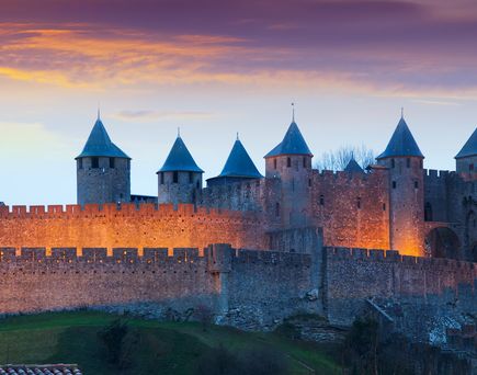 Eine imposante historische Burg oder Festung mit hohen Steinmauern und Türmen, beleuchtet von der untergehenden Sonne. Der Himmel ist in warmen Orangetönen gehalten.