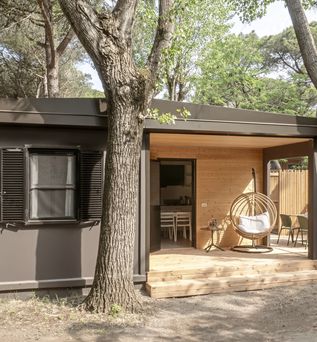 Modernes, naturfarbenes Mobilheim mit überdachter Holzveranda und Hängesessel, eingebettet in einen schattigen Pinienwald.