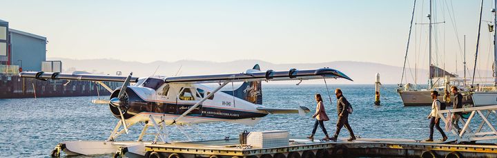  Ein Wasserflugzeug ist an einem schwimmenden Steg in einem Hafen festgemacht, während Passagiere an Bord gehen oder von Bord gehen. Im Hintergrund sind Segelboote und Gebäude am Ufer unter einem klaren Himmel zu sehen.