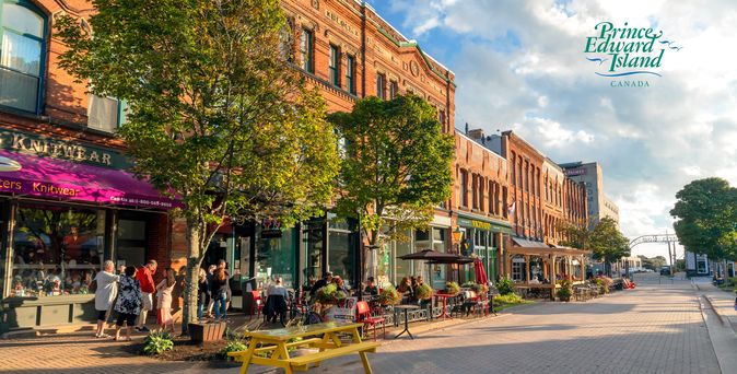 Zu sehen ist eine verkehrsberuhigte Straße in Charlottetown. Links einige braune Häuser, davor Cafes mit Sitzmöglichkeiten. Vor einem der Häuser ist eine rote Markise ausgefahren und es finden sich einige Bäume.
