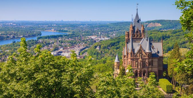 Eine imposante Außenansicht des Schlosses Drachenburg, das auf einem bewaldeten Hügel thront, mit seiner einzigartigen Architektur und einem Turm, der in den blauen Himmel ragt.