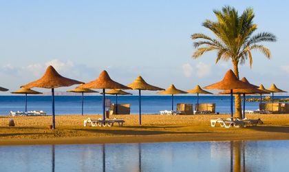 Ein Sandstrand mit Reihen von Strohsonnenschirmen und Liegestühlen, die sich an einem sonnigen Tag an einem Hotelresort in Hurghada, Ägypten, widerspiegeln.
