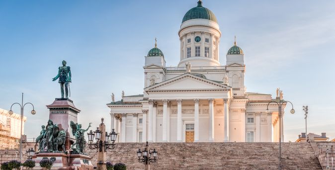 Die Helsinki Kathedrale auf einem erhöhten Platz mit breiten Steinstufen. Im Vordergrund befindet sich ein Denkmal mit einer Statue und einem Brunnen, umgeben von Bänken und Laternenpfählen, die den Senatsplatz in Helsinki beleuchten