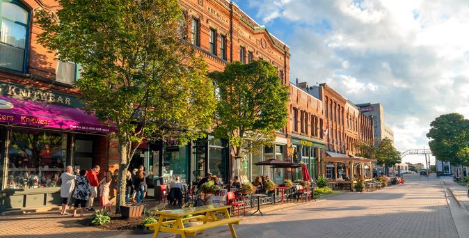 Zu sehen ist eine verkehrsberuhigte Straße in Charlottetown. Links einige braune Häuser, davor Cafes mit Sitzmöglichkeiten. Vor einem der Häuser ist eine rote Markise ausgefahren und es finden sich einige Bäume.