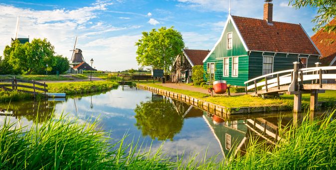 Ein kleines grünes Haus am Wasser mit einer Windmühle im Hintergrund.