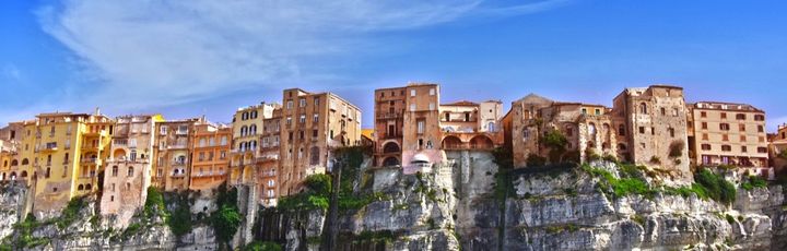 Die Stadt Tropea in Italien thront auf einer steilen Klippe über dem türkisfarbenen Meer, mit einem historischen Gebäude auf einem Felsen im Vordergrund.