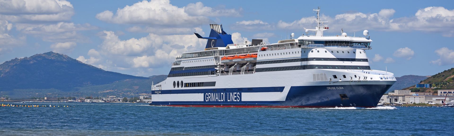 Eine Ansicht eines Grimaldi Lines-Schiffes, das auf dem offenen Meer fährt. Das Schiff ist weiß mit roten und blauen Details und hat den Schriftzug "GRIMALDI LINES" an der Seite. Der Himmel ist hell mit einigen Wolken, und das Wasser ist ruhig.