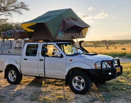Ein Nissan Navara Pickup-Truck mit einem auf dem Dach montierten Zelt steht auf einem Feldweg in einer weiten, offenen Landschaft.