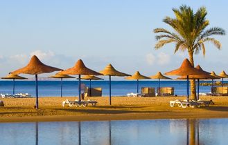 Ein Sandstrand mit Reihen von Strohsonnenschirmen und Liegestühlen, die sich an einem sonnigen Tag an einem Hotelresort in Hurghada, Ägypten, widerspiegeln.