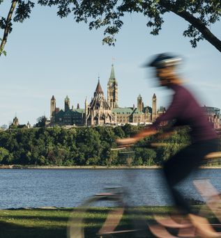 Fahrradfahrer fährt am Ottawa River entlang, im Hintergrund ist das Parlament zu sehen