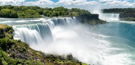 Zu sehen sind die beeindruckenden und weitläufigen Niagara Fälle, inmitten von hohen Bäumen. Blauer Himmel mit einigen kleinen weißen Wolken am Himmel.