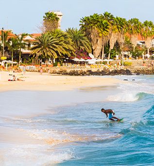 Surfer surft auf einer Welle im Hintergrund ein Dorf mit PAlmen