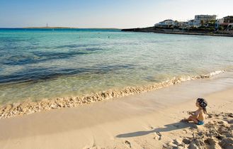 Ein Sandstrand mit Felsen und Meer, ein Baby sitzt im Sand mit blauem Kopftuch.