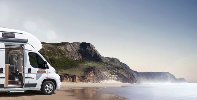 Ein modernes Weinsberg CaraTour 600 Wohnmobil steht auf einem Asphaltplatz, umgeben von Bäumen und einer grünen Landschaft unter einem bewölkten Himmel.