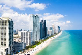 Blick auf Miami Beach. Links die hohen Hotels und Geschäftsgebäude, rechts daneben ein breiter weißer Sandstrand, daneben dann das türkisblaue Meer. Der Himmel strahlt hellblau.