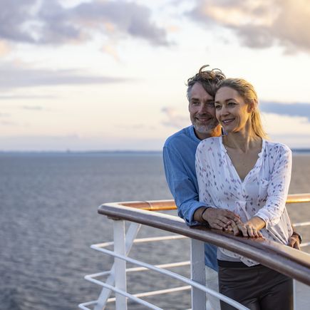 Eine Frau steht an der Reling eines Schiffes und blickt auf den Sonnenuntergang über dem Meer.