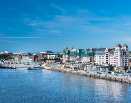Ein Kreuzfahrtschiff fährt in Oslo ein. Links das Schiff auf dem Meer, rechts die süßen netten Häuser direkt am Ufer. Blauer Himmel.