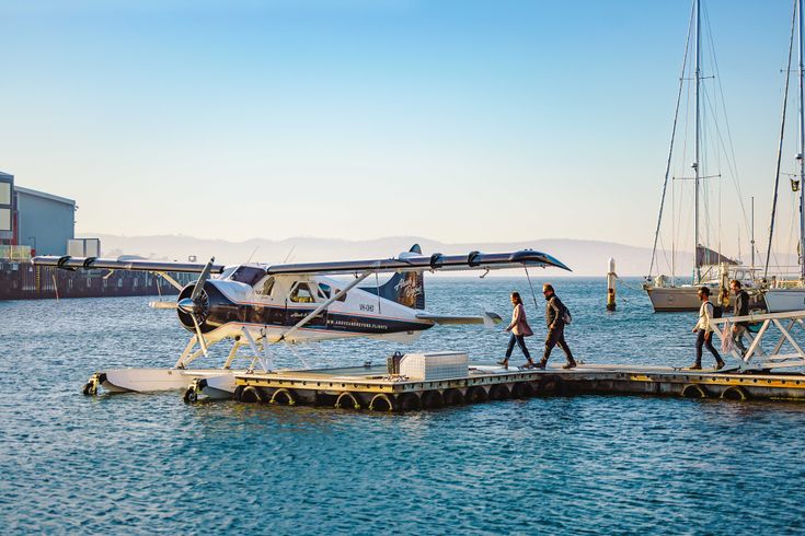  Ein Wasserflugzeug ist an einem schwimmenden Steg in einem Hafen festgemacht, während Passagiere an Bord gehen oder von Bord gehen. Im Hintergrund sind Segelboote und Gebäude am Ufer unter einem klaren Himmel zu sehen.