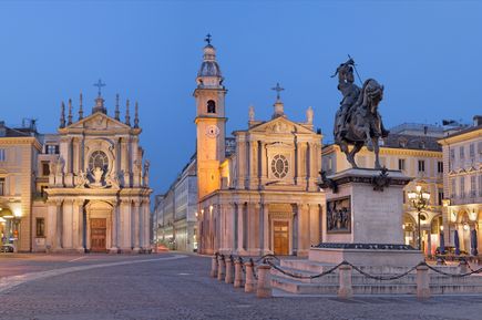 Blick auf den Markplatz Turins in der Dämmerung. Im Vordergrund eine Statur, dahinter ein Brunnen und viele schöne alte Gebäude. Eine Kirche ist ebenfalls im Hintergrund.