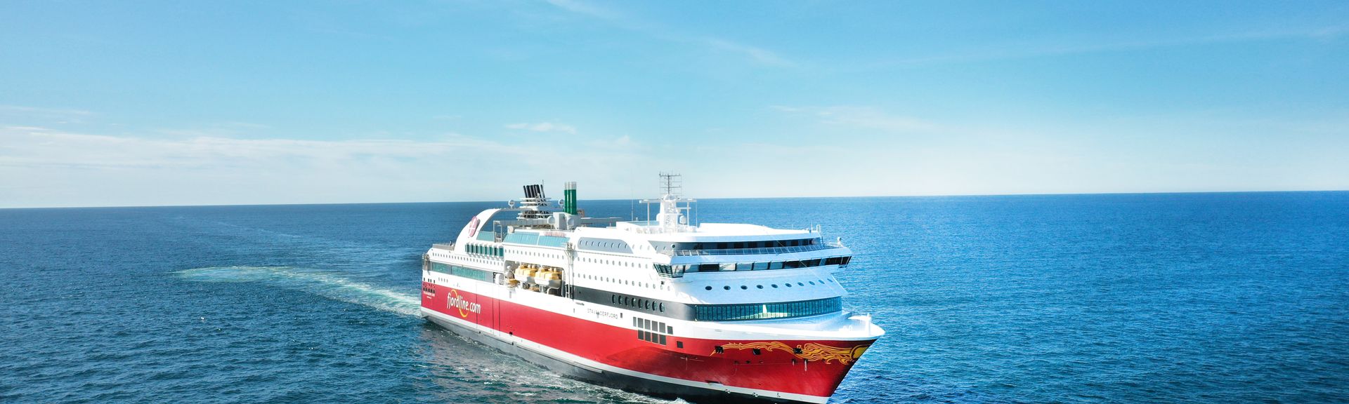 Ein großes rotes und weißes Fährschiff mit dem Namen "Fjord Line" an der Seite fährt auf dem offenen Meer, hinter sich eine Bugwelle ziehend, unter einem klaren blauen Himmel.