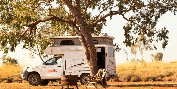  Ein weißer Apollo 4x4 Campervan steht unter einem schattenspendenden Baum in einer trockenen, von Gräsern bewachsenen Landschaft. Neben dem Fahrzeug sind ein Lagerfeuerring und ein Klappstuhl zu sehen.