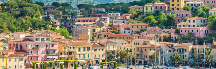 Blick auf Porto Azzuro vom Wasser aus. In der Front viele kleine Boote, dahinter viele schnuckelige bunte Häuser. Dahinter wiederum ein Berg und blauer Himmel.