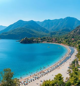  Panoramablick auf den Strand von Ölüdeniz, Türkei, mit zahlreichen Badegästen, türkisfarbenem Meer und bewaldeten Bergen unter blauem Himmel.