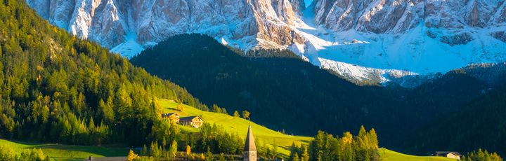 Die Dolomiten. Im Vordergrund grüne Wiese mit kleinem urigen Dorf, direkt am Waldhang. Dahinter die prächtigen, schneebedeckten Dolomiten bei blauem Himmel.