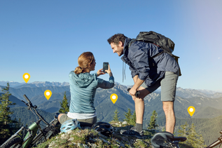 Ein Paar mit Mountainbikes macht auf einem Berggipfel eine Pause, die Frau fotografiert mit dem Handy die Aussicht, gelbe Standort-Markierungen sind in der Landschaft sichtbar.
