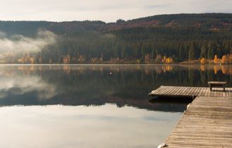 Ein ruhiger See mit einem Steg und Bäumen am Ufer, spiegelt den Himmel.