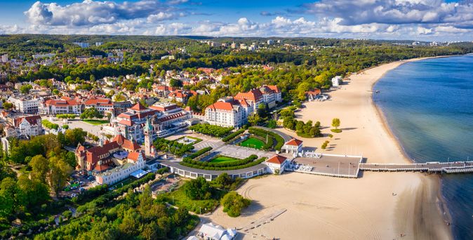 Blick auf Sopot. Links ist die Stadt zu sehen und vielen kleinen schönen Häusern. Von einem großen Haus führt ein großer breiter Steg ins Meer. Zwischen Wald und breitem Sandstrand.