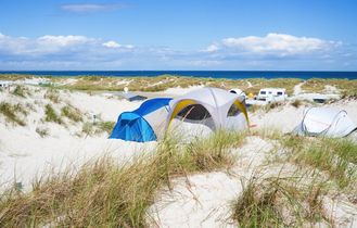 Blaues Zelt steht in den Sanddünen am Strand, dahinter Meer und blauer Himmel.