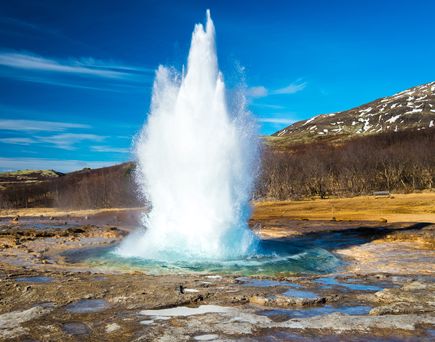 Blick über den lebendigen Geysir. Rundherum dürres Gras bzw. Landschaft. Im Hintergrund Berge und einige Bäume zu sehen. Blauer Himmel.