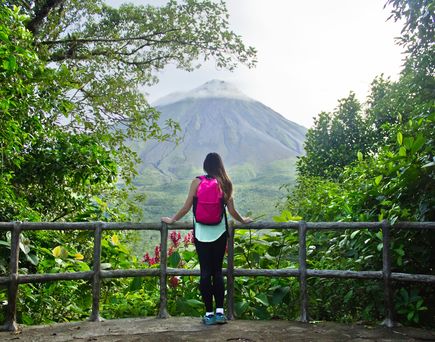 Touristin mit pinkem Rucksack am Rücken steht an Aussichtspunkt am Holzgeländer, Blick über die grüne Vegetation und den Berg im Hintergrund, blauer Himmel