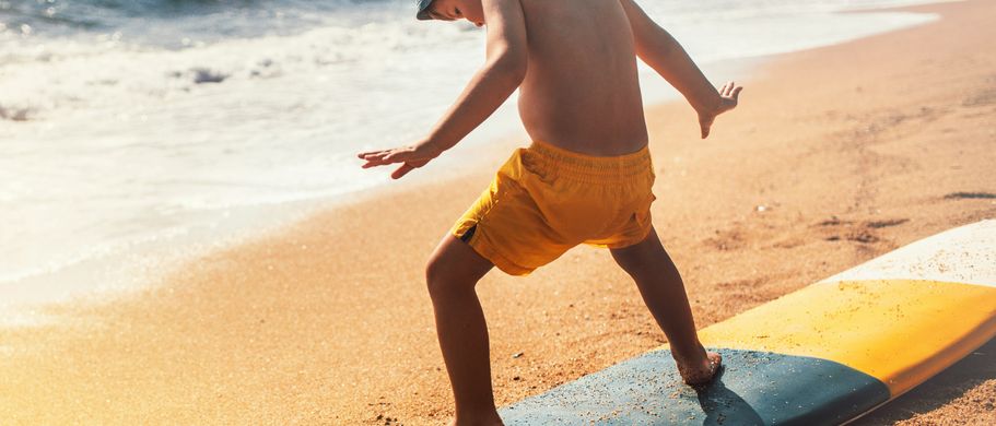 Ein kleiner Junge mit orangener Badehose steht auf einem Surfboard, welches noch am Starnd liegt.