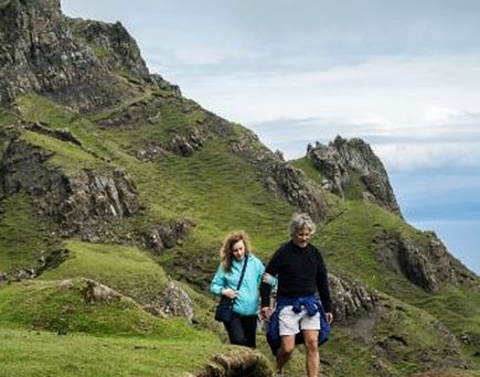Eine Person wandert auf einem Hügel mit Blick auf das Meer. Die Landschaft ist grün und felsig.