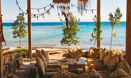 Eine gemütliche Strandbar in Dahab, Ägypten, mit schattenspendenden Strohdächern, Sitzgelegenheiten am Strand und Palmen mit Blick auf das Meer.